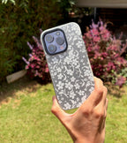 A hand holding a smartphone with a white and gray floral lace-patterned protective case, set against a blurred outdoor garden background.