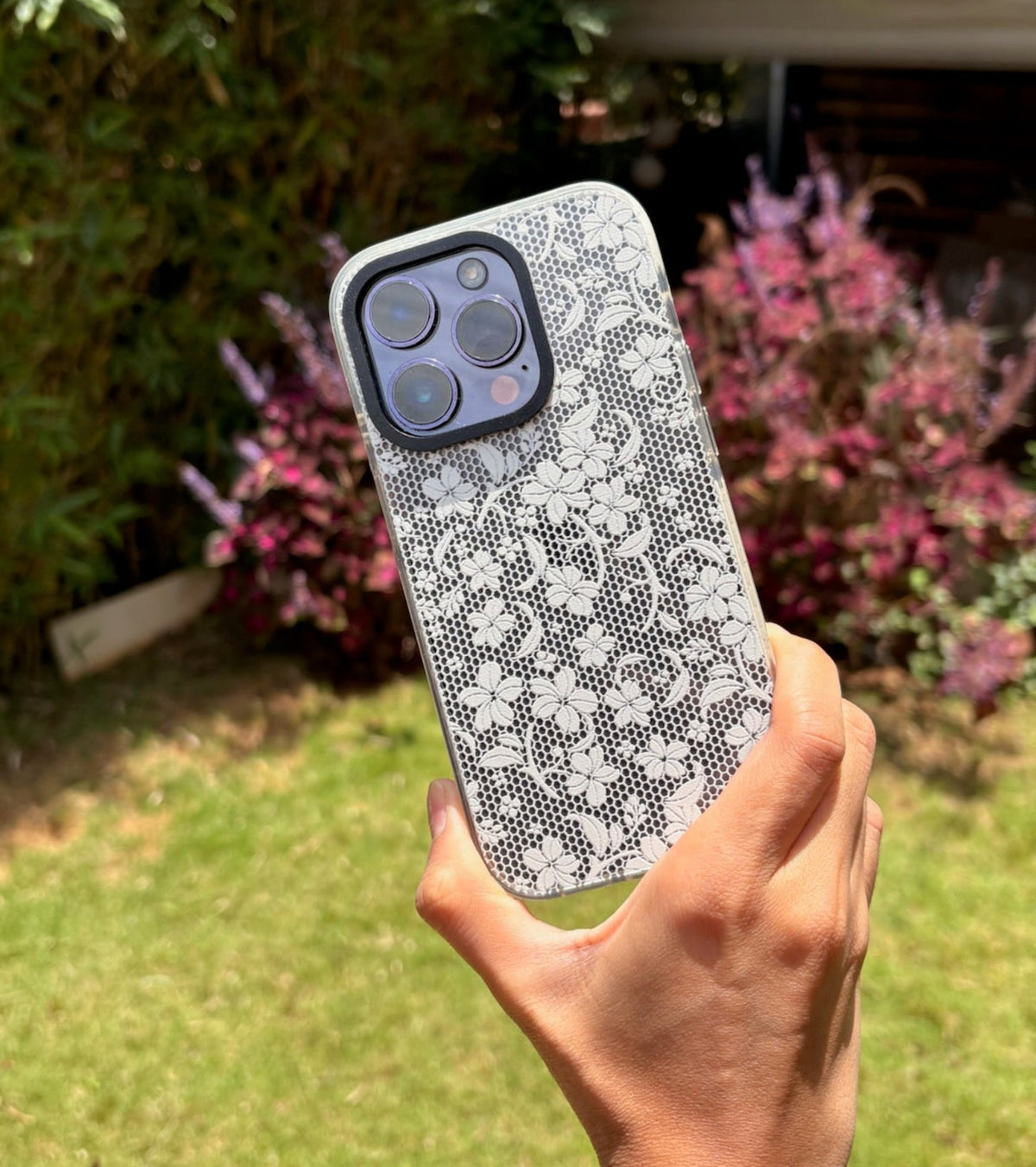 A hand holding a smartphone with a white and gray floral lace-patterned protective case, set against a blurred outdoor garden background.