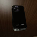 A black protective phone case with a silver metal edge, featuring the text 'This too shall pass' in white on the back, shown on a wooden surface.