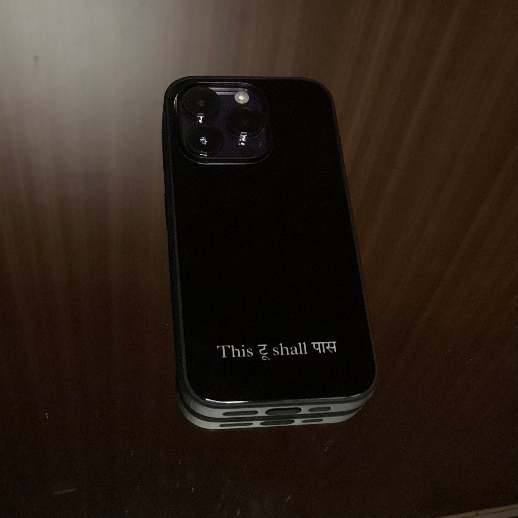 A black protective phone case with a silver metal edge, featuring the text 'This too shall pass' in white on the back, shown on a wooden surface.