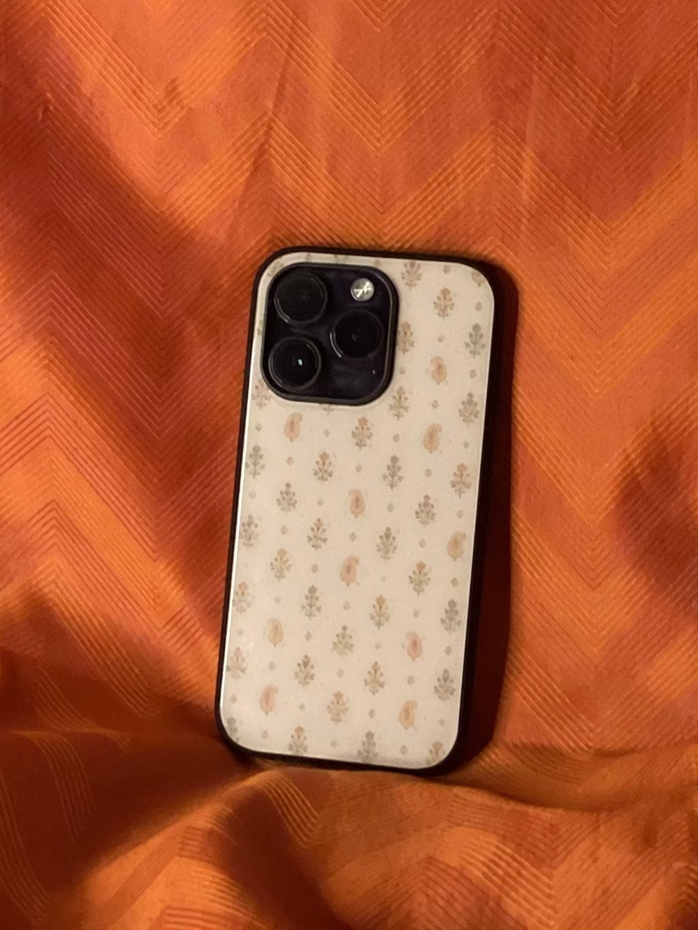 A phone case with a beige and brown floral pattern, shown on a black-edged smartphone, placed on a textured orange fabric background.
