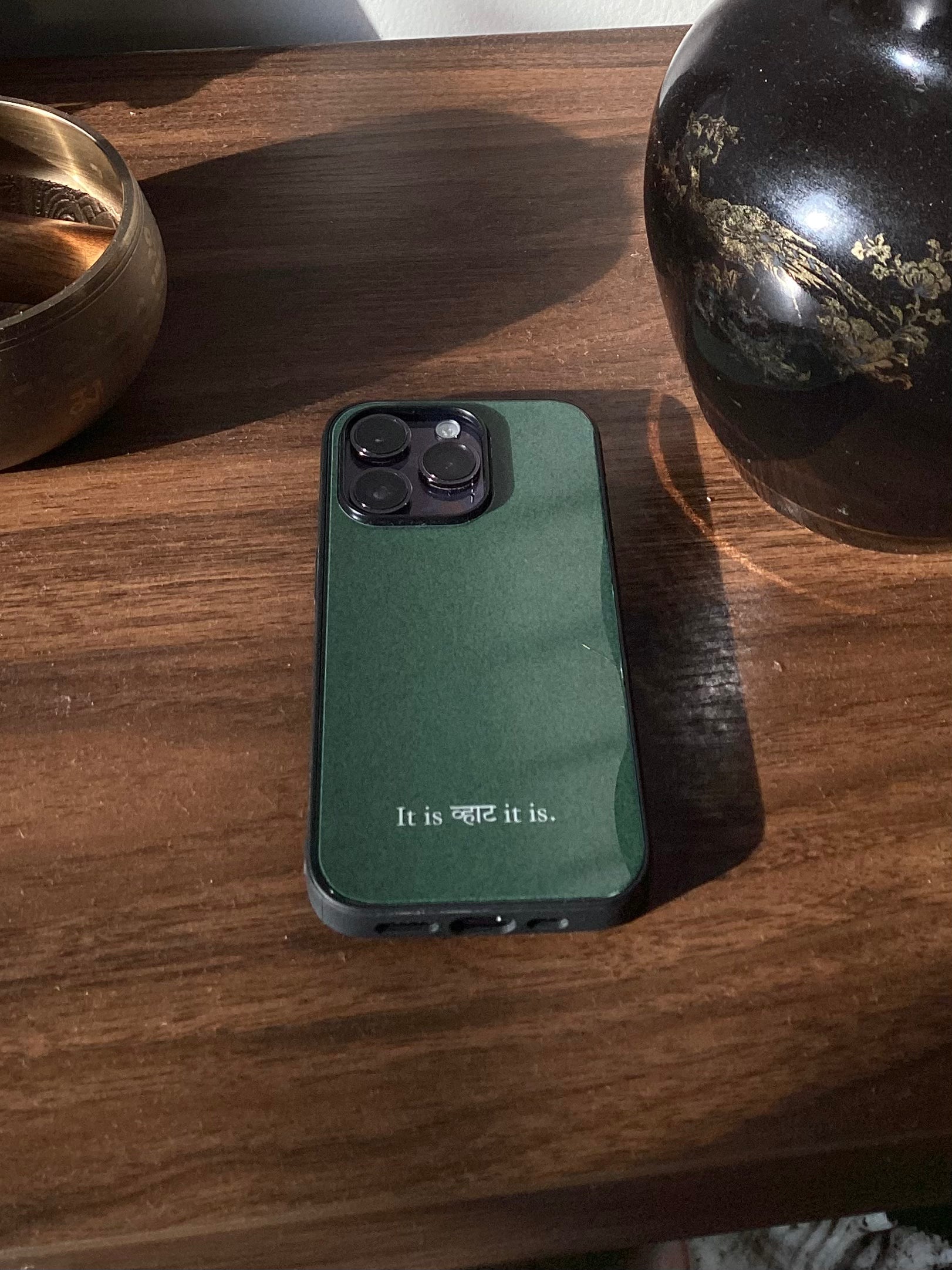 A dark green phone case with white text reading 'It is what it is.' resting on a dark wooden table, flanked by decorative ceramic vessels.