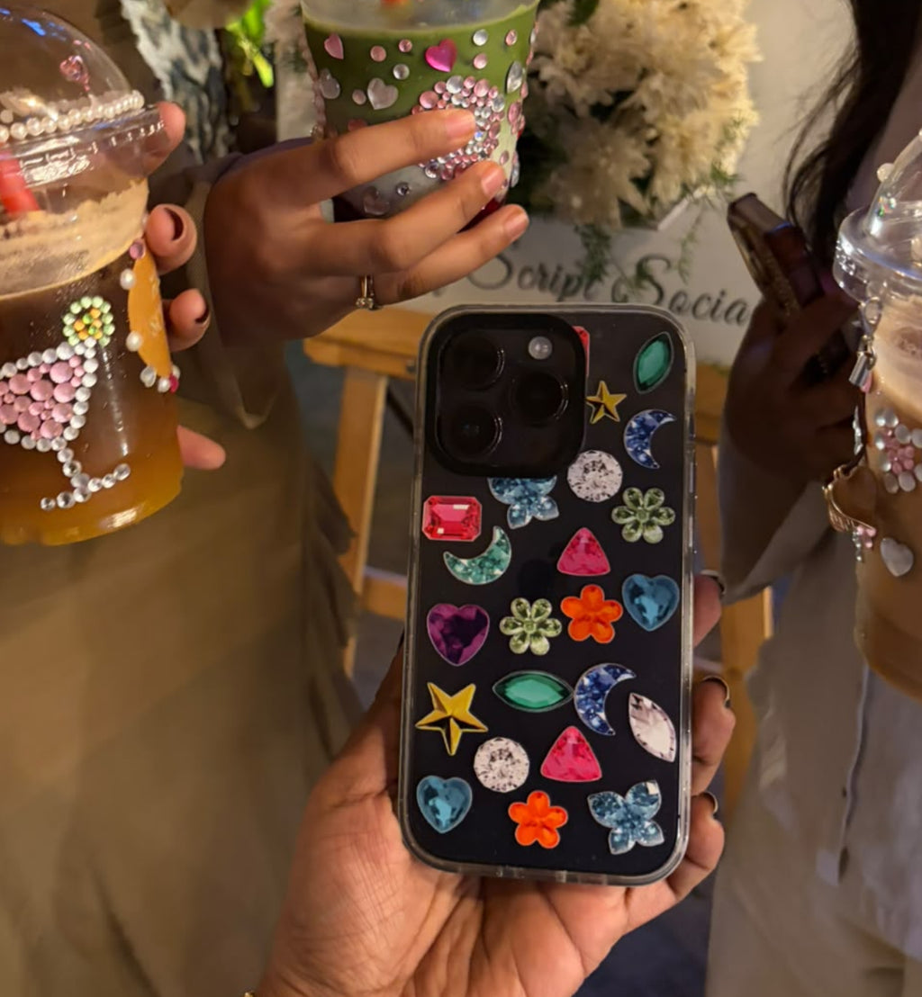 A person holds a black phone case decorated with colorful rhinestones and starfish motifs in the foreground. Two people holding decorated iced coffee drinks stand behind it in a social setting.