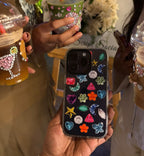 A person holds a black phone case decorated with colorful rhinestones and starfish motifs in the foreground. Two people holding decorated iced coffee drinks stand behind it in a social setting.