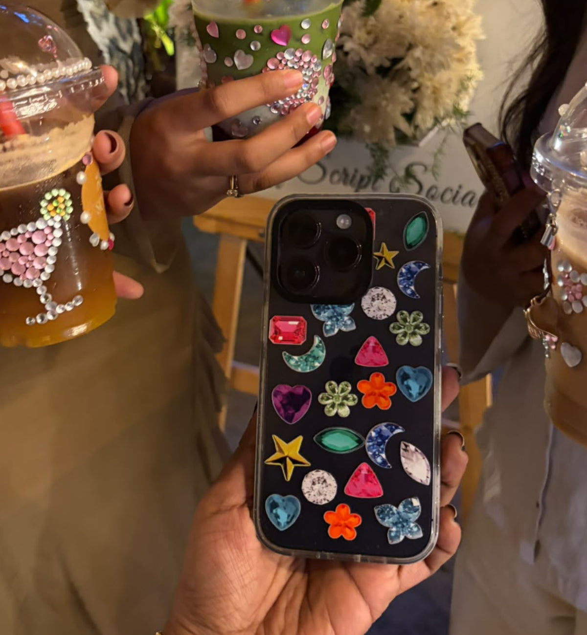A person holds a black phone case decorated with colorful rhinestones and starfish motifs in the foreground. Two people holding decorated iced coffee drinks stand behind it in a social setting.