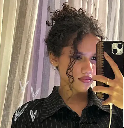 A woman taking a mirror selfie wearing a black pinstriped button-up shirt with white embroidered details on the shoulders. She has curly dark hair and is holding a black smartphone with a patterned case.