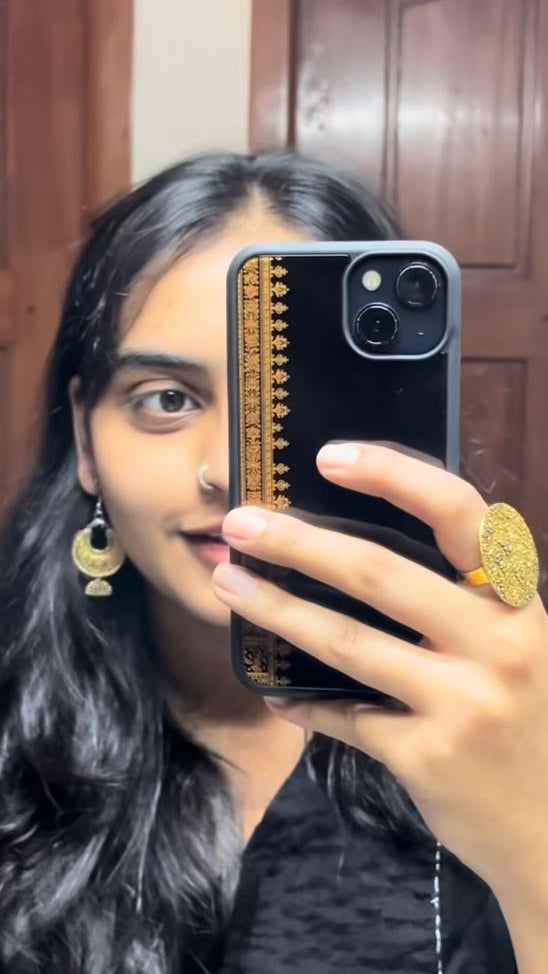 Person holding a phone with a mirror case, wearing gold earrings and a ring, in front of a wooden door.